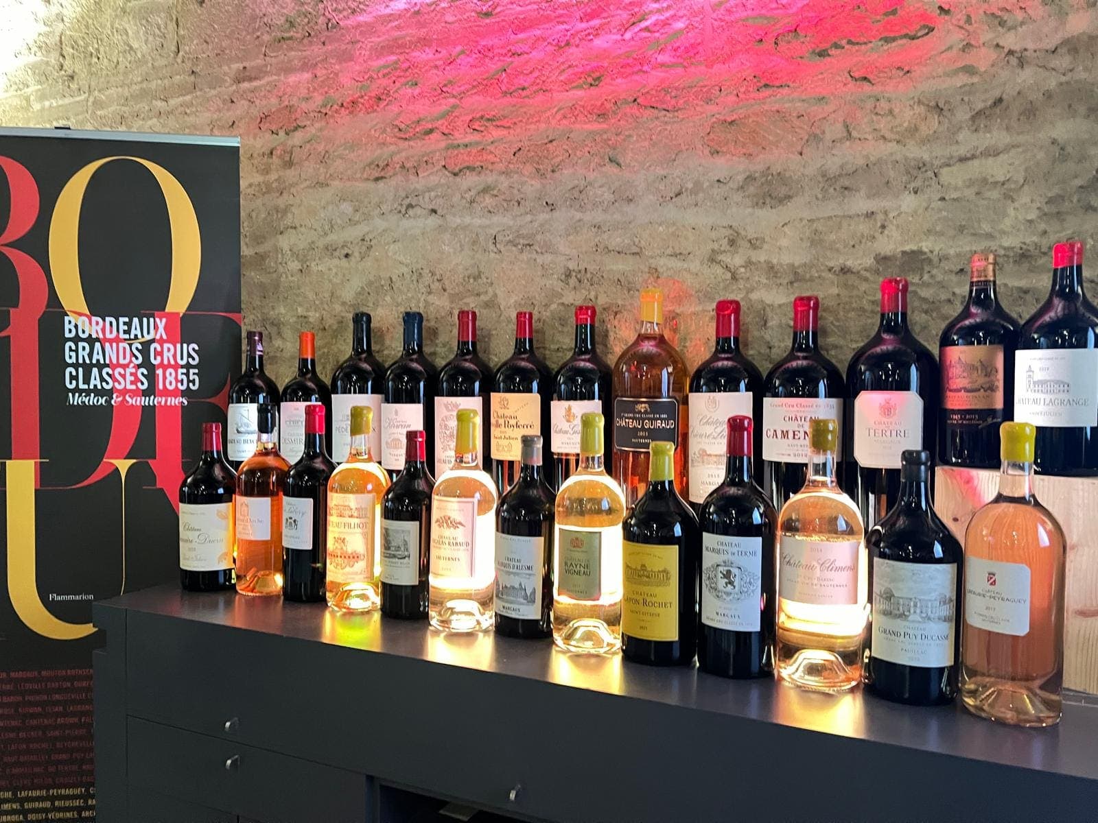 Grands formats de crus classés du Médoc et de Sauternes alignés sur un buffet, devant un mur de pierre éclairé de rose et une affiche « Bordeaux Grands Crus Classés 1855 »