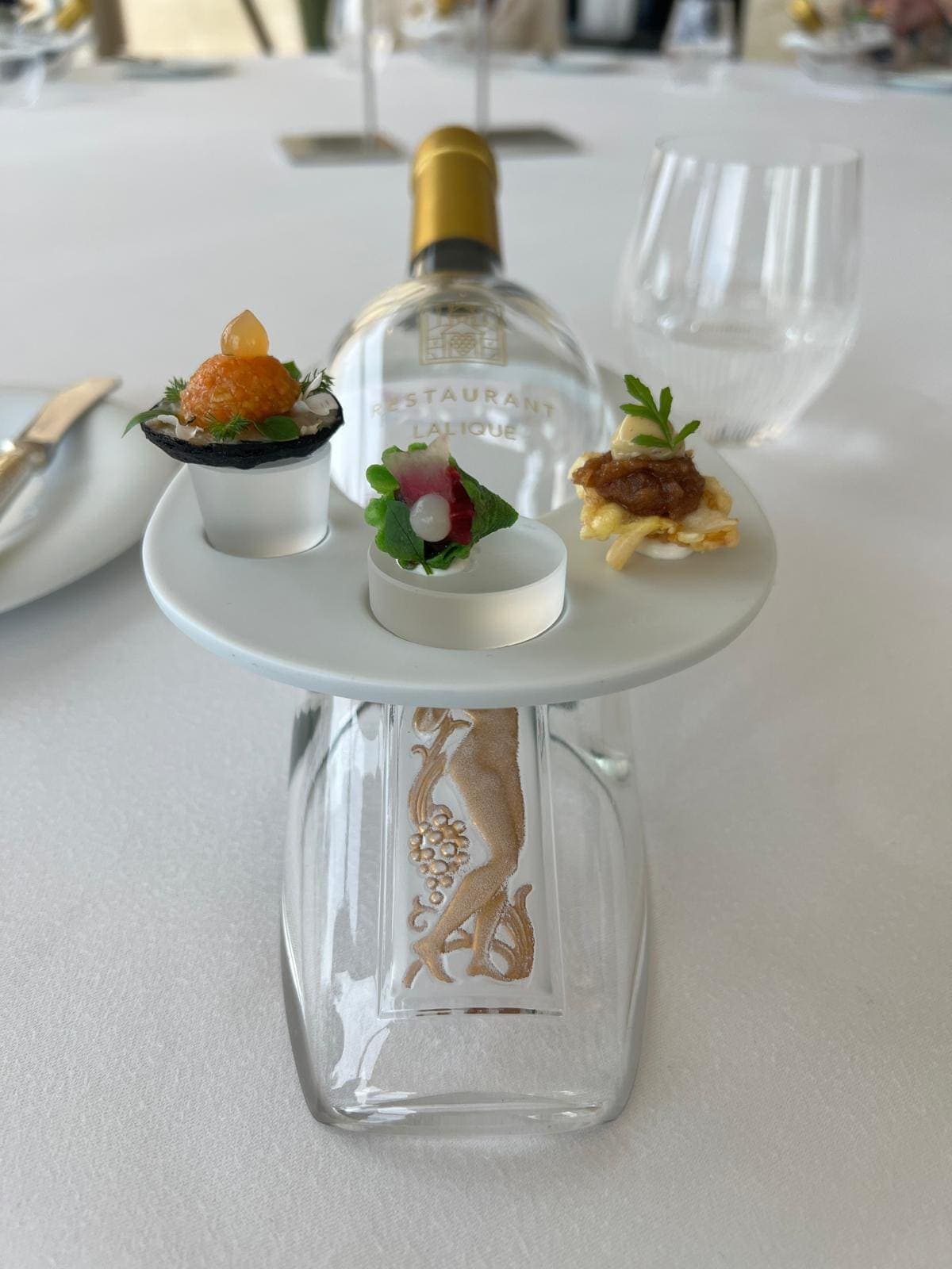 玻璃底座上呈现三道开胃小食,刻有"Restaurant Lalique"字样,背景为水瓶