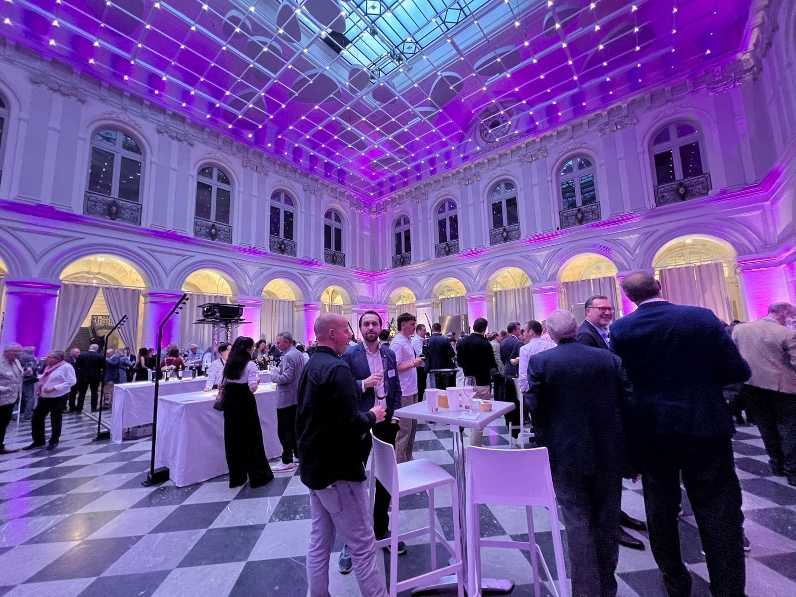 Réception debout dans une cour à arcades sous verrière illuminée de pourpre, sol en damier, invités autour de tables hautes
