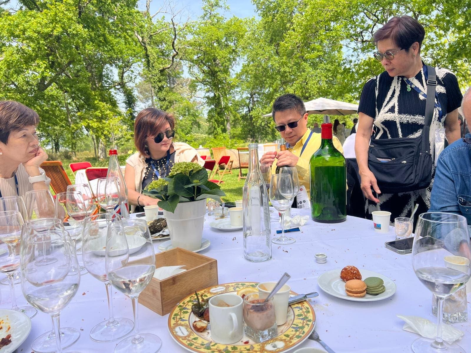Groupe de convives autour d’une table dressée en extérieur, sous de grands arbres