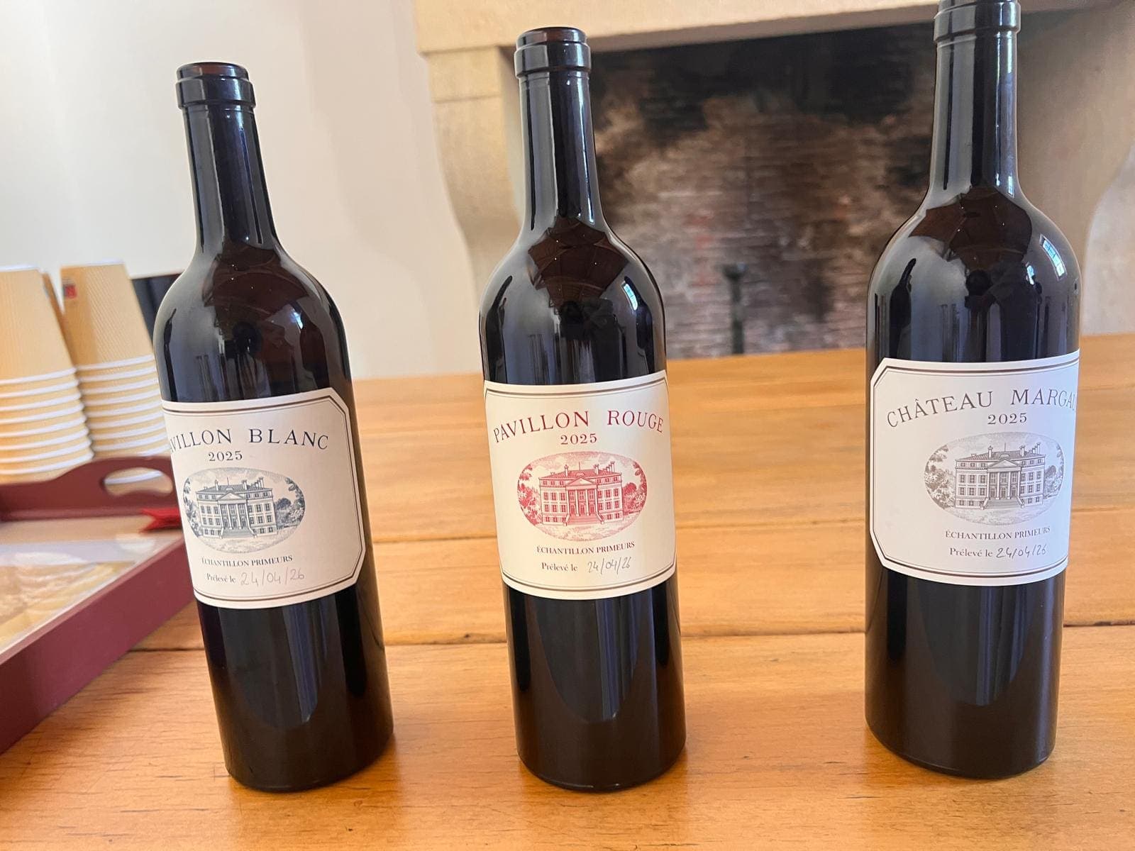 Trois bouteilles d’échantillon primeurs Château Margaux (Pavillon Blanc, Pavillon Rouge, Château Margaux) alignées devant une cheminée
