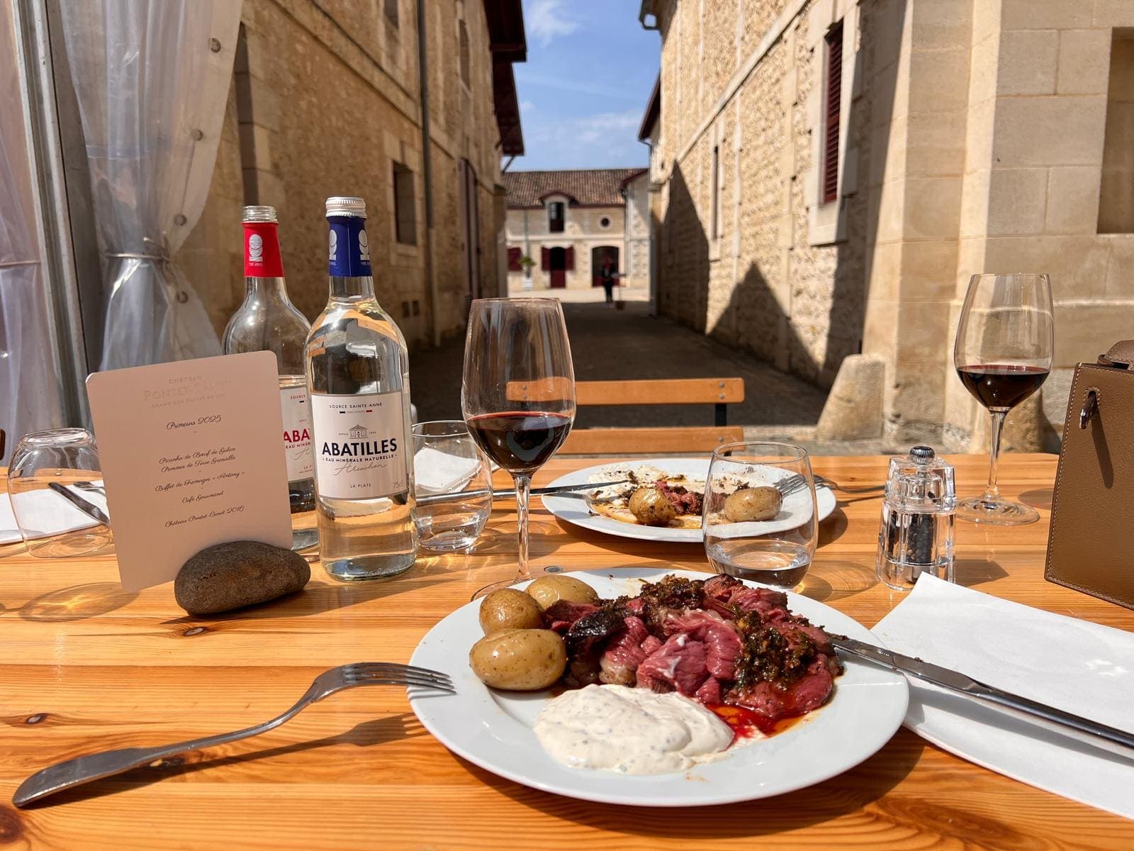 Déjeuner en terrasse entre bâtiments en pierre d’un château, assiette de viande et verres de vin