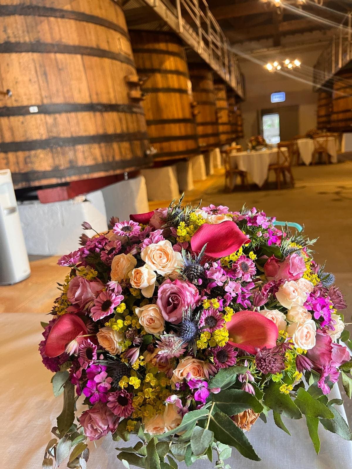Bouquet de fleurs roses et blanches devant des foudres en bois, tables dressées en fond dans un chai