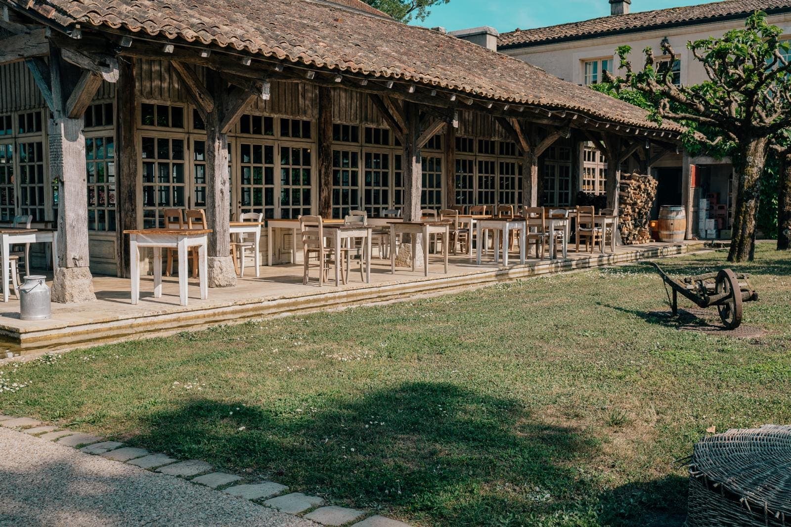 Terrasse couverte d’un domaine bordelais, tables en bois, charrette ancienne et jardin