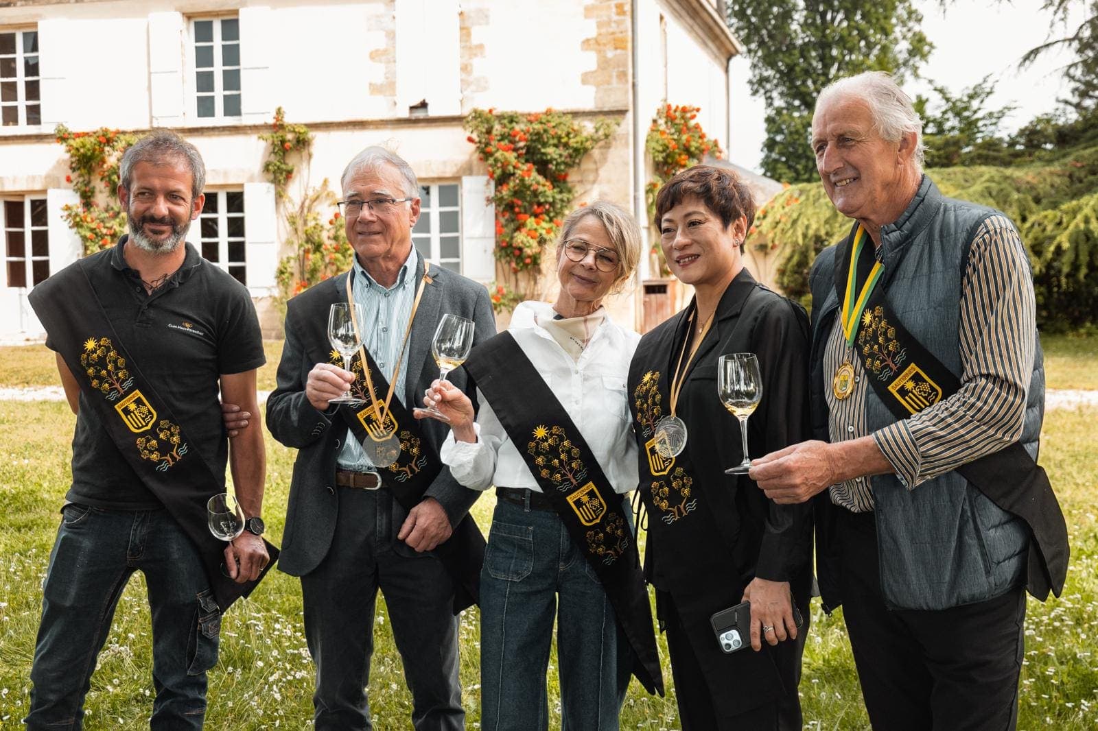Groupe de cinq personnes portant l’écharpe d’une confrérie viticole du Sauternais, verres à la main, devant un château fleuri