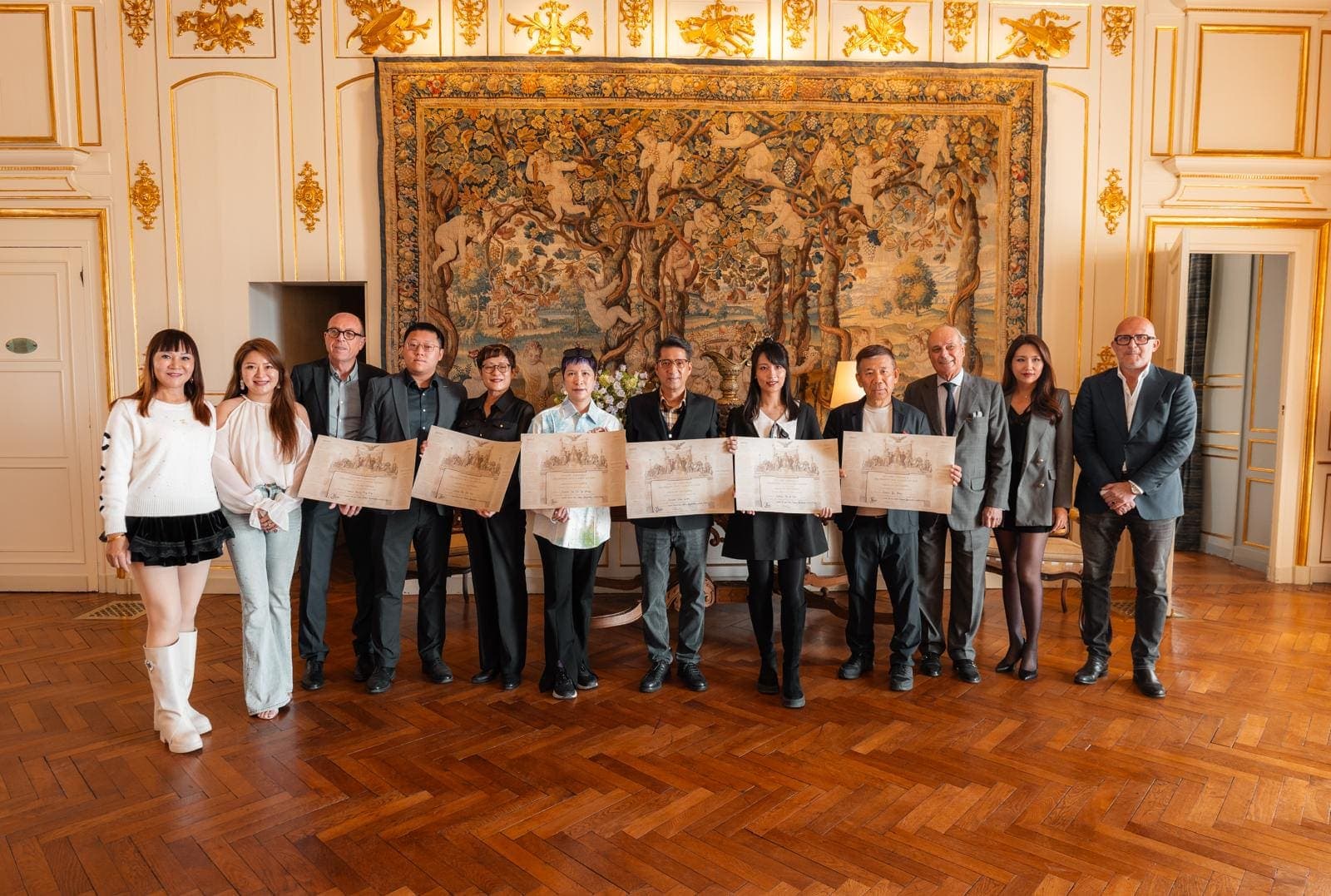 Groupe d’une douzaine de personnes tenant des diplômes, devant une grande tapisserie ancienne dans un salon doré