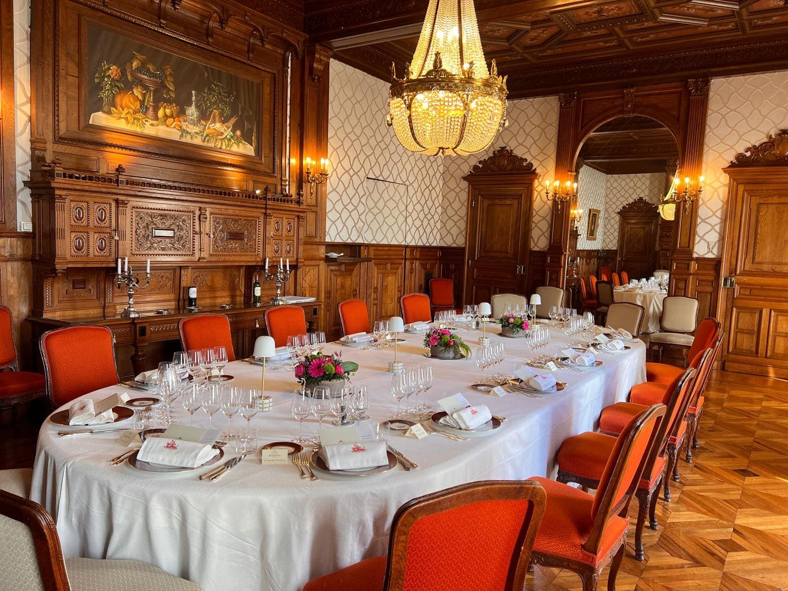 Salle à manger de château aux boiseries sombres, chaises orange et grand lustre en cristal, table dressée pour un dîner