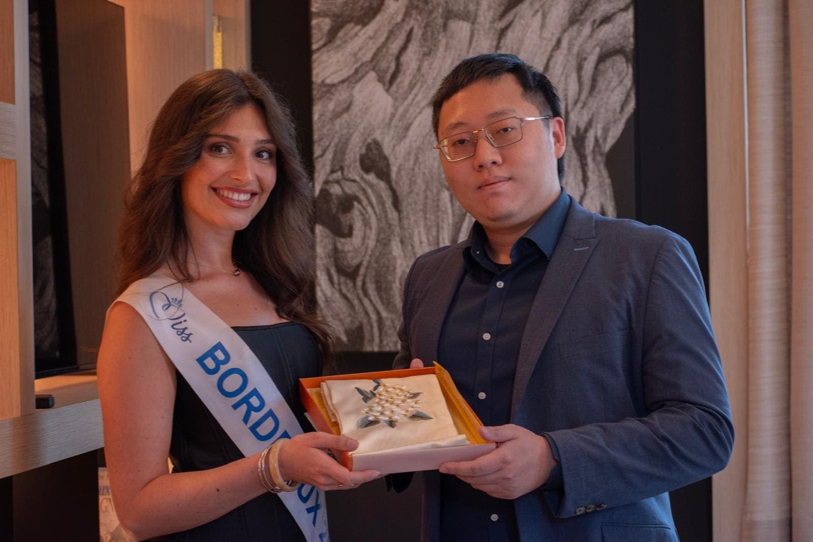Miss Bordeaux, écharpe portée, remet un présent brodé à un partenaire lors d’une réception