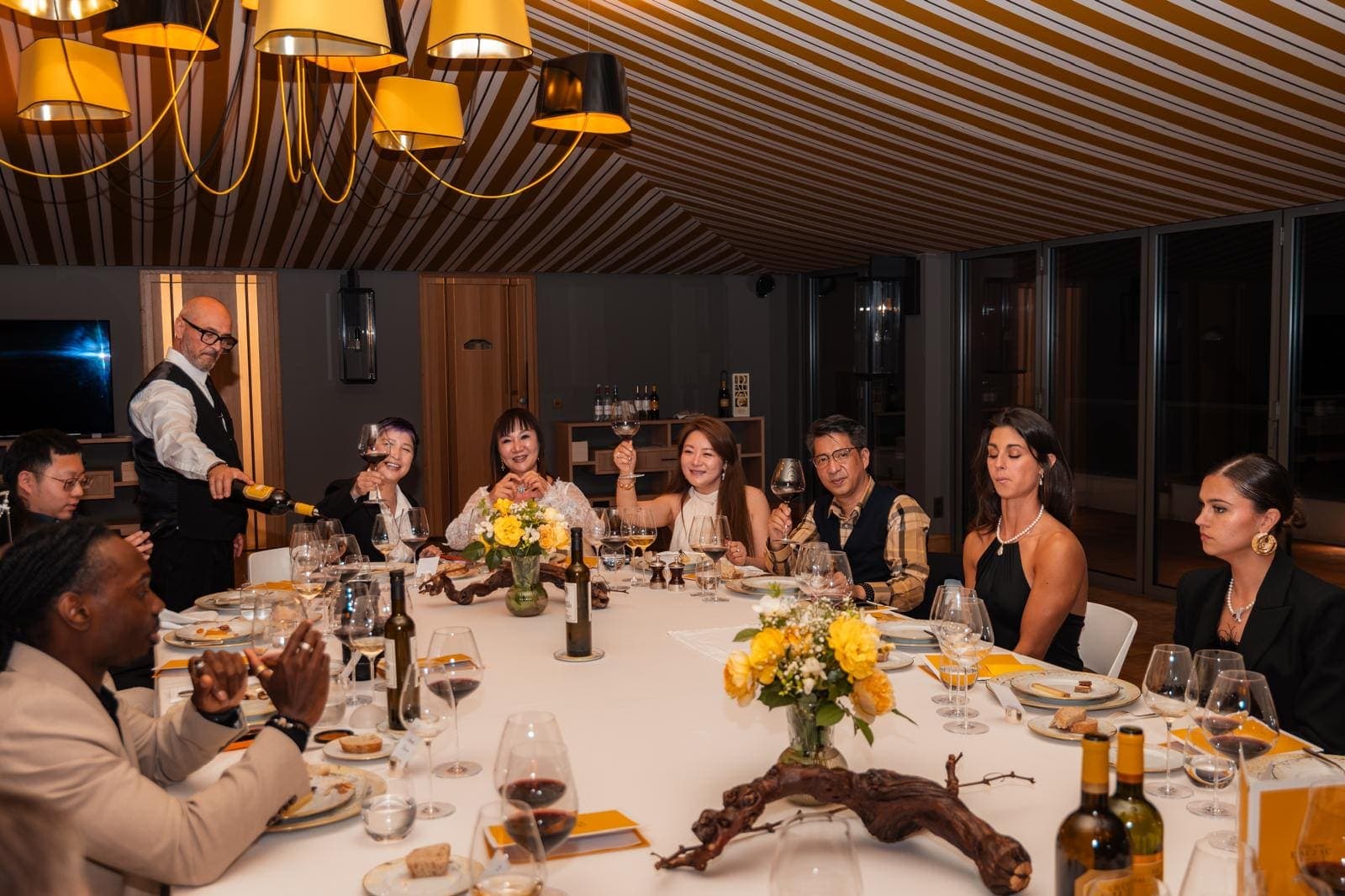 Convives portant un toast autour d’une table dressée, sommelier servant le vin, dîner sous tente